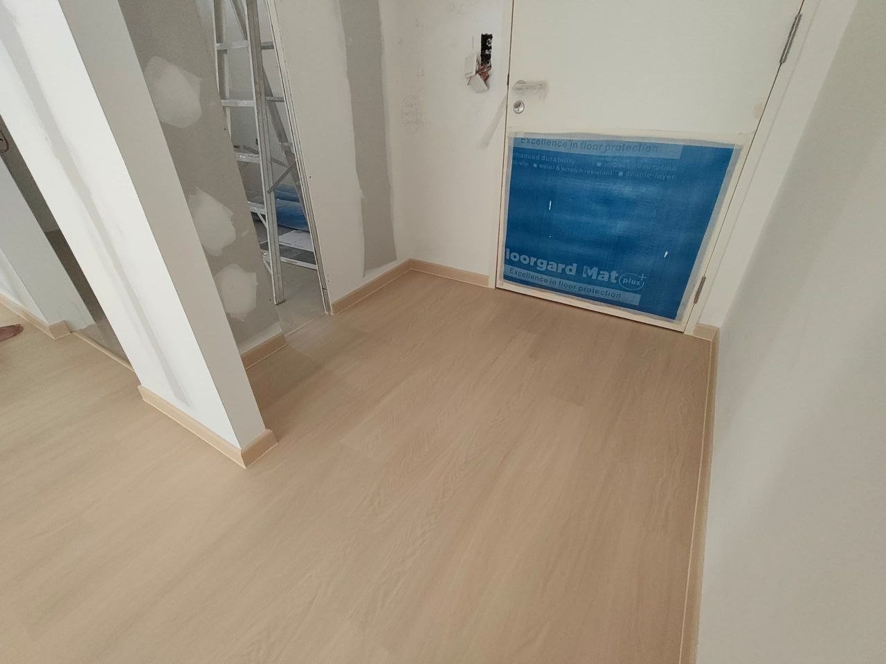 Light oak-look SPC floor meeting matching skirting in a corner, with a blue floor-protection mat on the door, in a D'Clover Residences unit